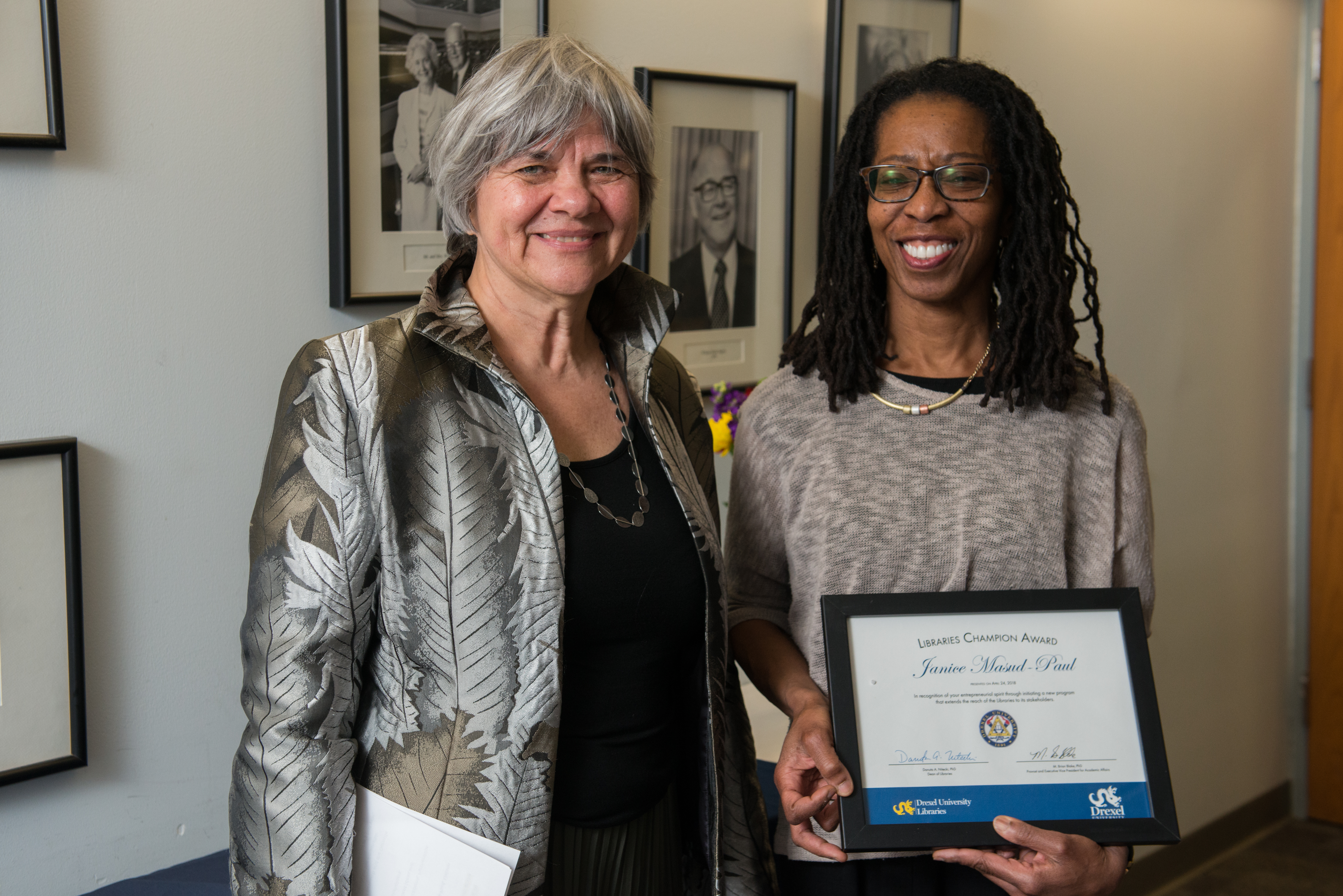 Two women smiling, face the camera. The woman on the right side holds a framed award.